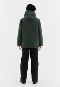 Groene waterdichte jas met capuchon, gestructureerde stof en zijzakken, gedragen met zwarte broek en een zwarte gebreide beanie.
