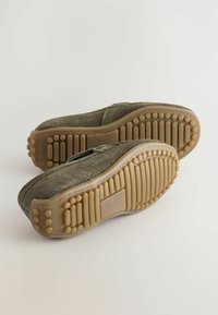 Des chaussures à enfiler en daim vert avec des semelles en caoutchouc texturé présentant des motifs ronds et striés pour une meilleure adhérence. Design minimaliste sans logos visibles.