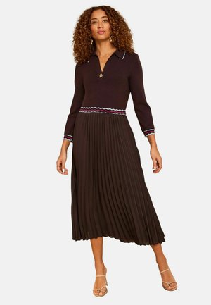 Žena s kučeravými vlasmi, oblečená v tmavohnedých šatách s dlhým rukávom, plisovanou sukňou a pruhovaným lemom v páse a na manžetách, stojaca s nohami od seba.