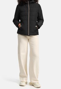 Osoba oblečená v černé zateplené bundě s zipem, smetanových širokých kalhotách a světle zbarvených masivních teniskách, stojící s rukama v kapsách.