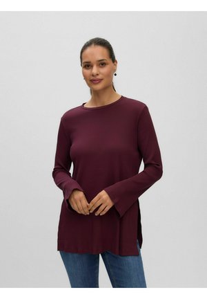 Femme aux cheveux foncés attachés, vêtue d'une chemise bordeaux à manches longues et d'un jean bleu, se tenant devant un fond clair uni.