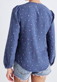 Femme portant un chemisier bleu à manches longues avec de petites fleurs roses et blanches, associé à un short en jean bleu clair effiloché, vue de dos.