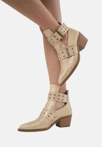 Botines de cuero beige hasta el tobillo con punta afilada, correas con hebilla y tachuelas decorativas. Presenta un tacón de madera de altura media.