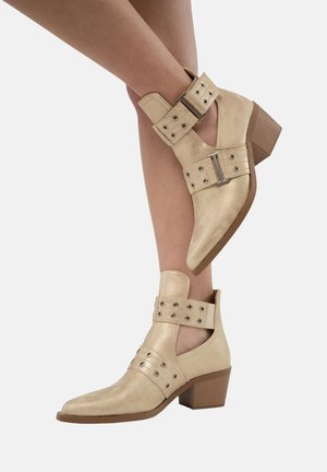 Botines de cuero beige hasta el tobillo con punta afilada, correas con hebilla y tachuelas decorativas. Presenta un tacón de madera de altura media.