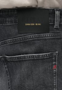 Μαύρα τζιν με μαύρη ετικέτα που φέρει την ένδειξη "REPLAY DNM GDS 1981." Διαθέτει κόκκινο κεντητό λογότυπο στην πίσω τσέπη και ορατές λεπτομέρειες ραψίματος.