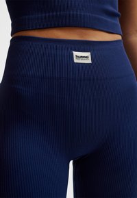 Marinblå ribbstickade leggings med hög midja; har en Hummel Performance-logotyp nära toppen. Tyget verkar stretchigt och texturerat.