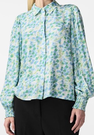 Langærmet bluse med grønt, blåt og hvidt abstrakt mønster, knaplukning foran, puffede ærmer og smockede manchetter, båret med sorte bukser.