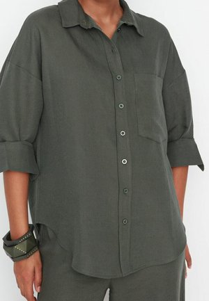 Kvinde iført løs, mørkegrøn skjorte med knapper og opkørte ærmer, matchende bukser og et chunky mørkegrønt armbånd på venstre håndled.