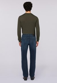 Hombre de espaldas vestido con una camisa de manga larga verde oliva, jeans rectos azul oscuro y zapatos negros sobre un fondo liso.