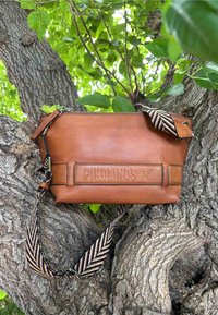 Bolso de piel marrón con el logotipo "Pikolinos" en relieve, que presenta una correa de diseño y cierre con cremallera. Colocado sobre el tronco de un árbol texturizado.