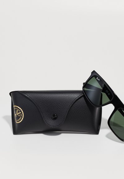 Черен чифт слънчеви очила Ray-Ban с зелени стъкла, поставени до черен текстуриран твърд калъф с золотист и черен логотип.