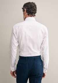 Camicia bianca a maniche lunghe, con colletto classico e polsini con bottoni, abbinata a pantaloni blu scuro a righe sottili.