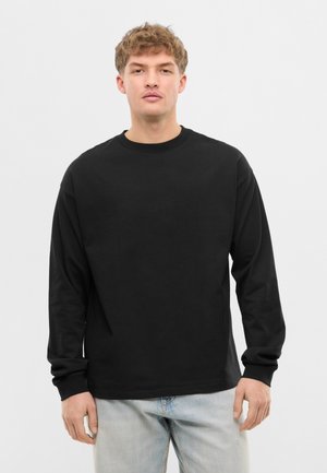 Jonge man met kort krullend haar, draagt een effen zwart shirt met lange mouwen en lichtblauwe spijkerbroek, staand tegen een neutrale achtergrond.