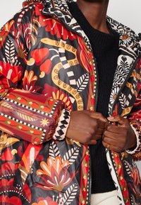 Chaqueta acolchada con un vibrante estampado floral y abstracto en rojo, naranja y negro. Cuenta con un cierre de cremallera y puños elásticos.