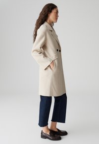 Beige wollen blend jas met een notch-kraag, dubbele knoopsluiting, zijzakken en een relaxte pasvorm. Gedragen met een donkere spijkerbroek en loafers.