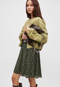 Khakifarbene Bomberjacke mit elastischen Bündchen, grünes Blumenmusterkleid mit ausgestelltem Rock, hält eine braune glänzende Handtasche mit Kettenakzenten.