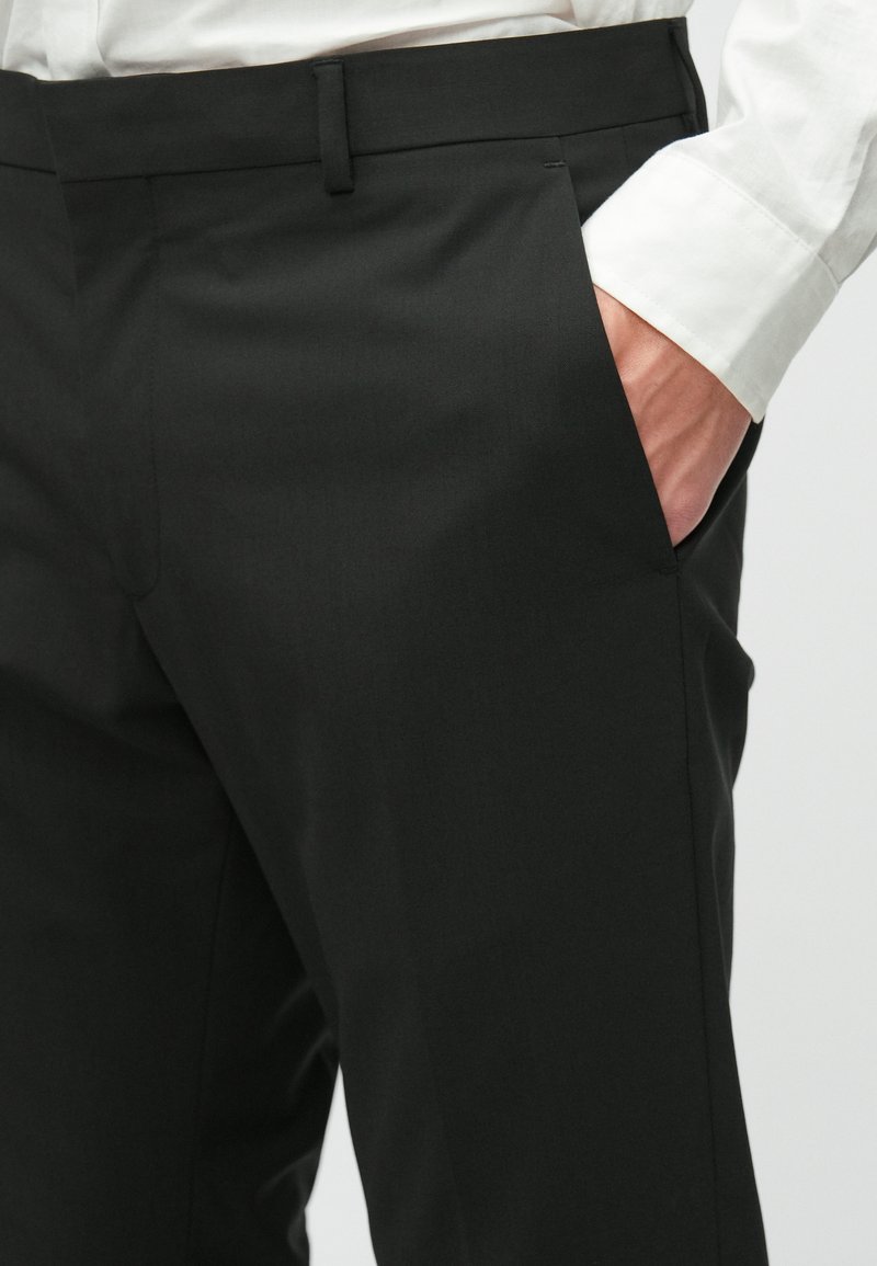 Schwarze maßgeschneiderte Hose mit glatter Textur, ausgestattet mit einer Vordertasche und einem schlanken Schnitt, kombiniert mit einem weißen Hemdsärmel.