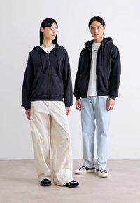 Felpe nere con zip indossate da due modelli. Uno indossa pantaloni relaxed-fit color avorio, l'altro jeans azzurri chiari. Entrambi portano sneakers nere. Design minimalista.