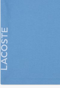 Ανοιχτό μπλε βαμβακερό T-shirt με τη λέξη "LACOSTE" εκτυπωμένη κάθετα με λευκά γράμματα στην αριστερή πλευρά, με ομαλή, ομοιόμορφη υφή.