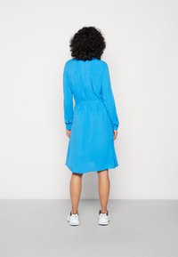 Abito camicia blu con maniche lunghe, vita cintrata e orlo al ginocchio. Realizzato in un tessuto liscio. Indossato con sneakers bianche.