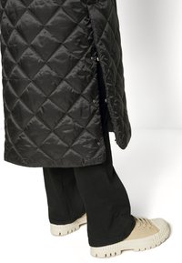 Abrigo acolchado negro con acabado brillante, que cuenta con cierres a presión a los lados y se combina con pantalones oscuros y zapatillas beige.