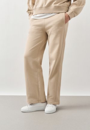 Άτομο που φοράει μπεζ φόρμα και αντίστοιχο φούτερ με λευκά αθλητικά, στέκεται μπροστά σε απλό ανοιχτό γκρι φόντο.