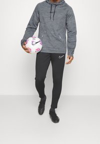 Szara bluza z kapturem o teksturowanym wzorze, czarne legginsy sportowe z białym logo oraz czarne korki piłkarskie, trzymając wzorzystą piłkę nożną.