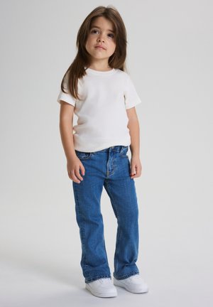 Nuori lapsi, jolla on pitkät ruskeat hiukset, valkoinen lyhythihainen paita, siniset farkut ja valkoiset tennarit, seisoo vaaleaa taustaa vasten.