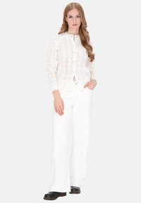 Cardigan blanc en tricot avec un motif géométrique, encolure ronde, fermeture à boutons, associé à un pantalon blanc à jambes larges et des chaussures noires.