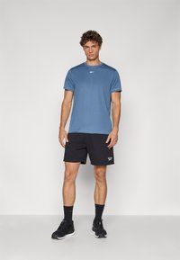 Blå atletisk t-shirt med korte ærmer, logo på brystet, parret med sorte shorts. Sorte atletiske sko og mørke sokker fuldender outfittet.