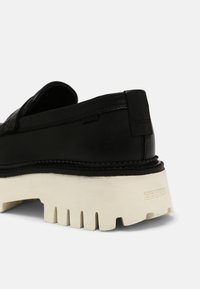 Mocassin en cuir noir avec une semelle épaisse et chunky blanche dotée de profondeurs, marqué "BRONX" sur le talon et sur le côté près de la couture.