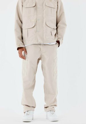 Chaqueta de utilidades beige con múltiples bolsillos frontales y botones a presión, combinada con pantalones cargo beige a juego. Ambos están hechos de un tejido resistente.