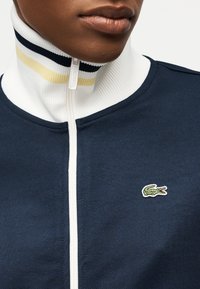 Κοντινό πλάνο ενός ατόμου που φοράει μπλε σκούρο φούτερ με φερμουάρ, με λευκή γιακά ριγέ σε μπλε και μπεζ, με μικρό πράσινο λογότυπο κροκόδειλου.