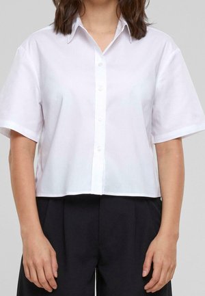 Woman wearing a short-sleeve white button-up shirt tucked into loose black pants, standing against a plain background.