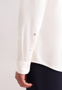Witte lange mouwen overhemdenmanchet met knopen en een klein zwart geborduurd bloemdetail op de mouw, gedragen met donkere broek.