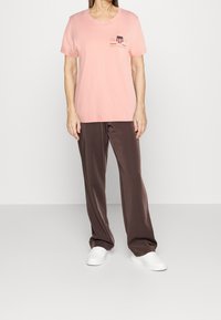 T-shirt en coton rose clair avec des manches courtes et un logo graphique sur la poitrine, associé à un pantalon en satin marron foncé et des baskets blanches.