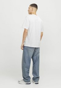 Hvid bomulds T-shirt med korte ærmer, parret med løstsiddende blå denim jeans. Modellen vender væk fra kameraet, hvilket afslører det enkle design.