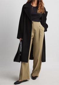 Černý vlněný kabát se širokým límcem, doplněný volnými olivově zelenými kalhotami s vysokým pasem. Model drží černou kabelku.