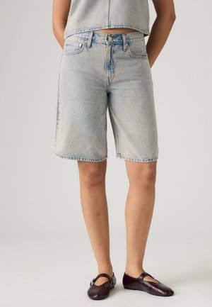Person trägt hellblaue Denim-Bermuda-Shorts, passendes kurzes Denim-Top und dunkelburgunderfarbene Mary Jane Ballerinas, steht vor einem einfarbigen Hintergrund.