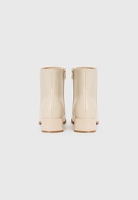 Bottines beiges en matériau synthétique brillant. Elles possèdent un talon carré et une fermeture éclair sur le côté. Texture lisse et design minimaliste.