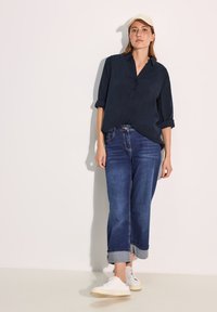 Camicia blu navy con maniche arrotolate, abbinata a jeans in denim a gamba larga con orlo risvoltato e sneakers bianche. Outfit casual.