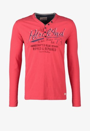 Rotes Langarmshirt mit Henley-Ausschnitt und Druckdetail, kombiniert mit grauer Jeans und weißen Sneakers, dazu eine braune Umhängetasche.