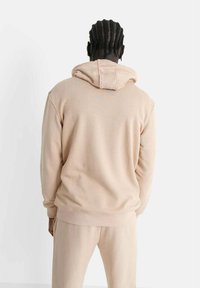 Sudadera beige con capucha y puños acanalados, fabricada en material suave. Presenta un sutil detalle de logo en la parte posterior. Complementa pantalones jogger a juego.