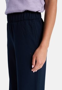 Mörkblå träningsbyxor med elastisk midja, sidofickor och en liten logotypdetalj på vänstra låret, tillverkade av mjukt tyg.