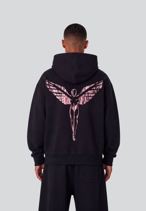 Man draagt een zwarte hoodie met een glanzende roze gestileerde gevleugelde figuur op de rug, staand tegen een effen grijze achtergrond.