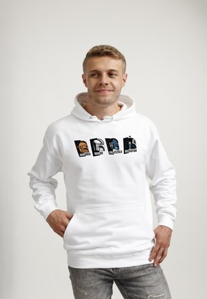 Ung mand med kort hår iført en hvid hættetrøje med Star Wars Mandalorian karakter hjelme og navne, stående med hænderne i lommerne.