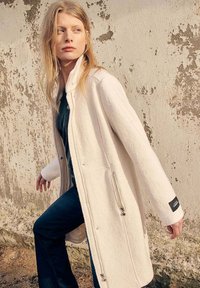 Manteau blanc en tissu texturé, semblable à de la polaire, avec un col montant et une fermeture éclair à l'avant. Le modèle l'associe avec un jean foncé.