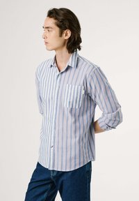 Camisa de botones a rayas en azul y blanco con acentos rojos; cuenta con un bolsillo en el pecho, mangas largas arremangadas hasta los codos y un dobladillo curvado.