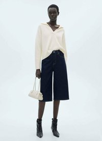 Romige geribde trui met een v-hals, gecombineerd met donkere denim culottes. Het model houdt een kleine romige tas vast en draagt zwarte hoge hakken.