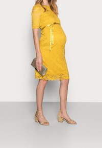 Gul spetsklänning med korta ärmar, vågig nederkant och satinband. Matchad med beige blockklacksandaler och en silverfärgad kuvertväska.